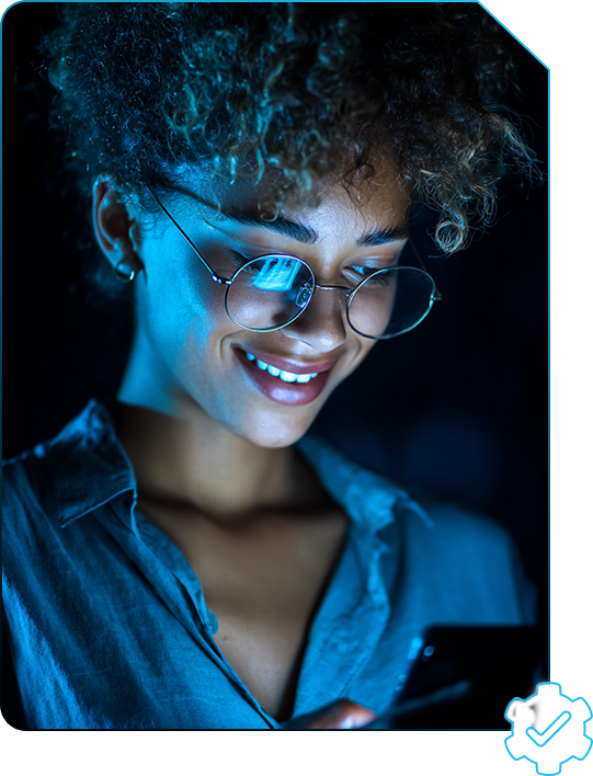 Mulher negra de cabelo cacheado usando óculos redondo e camiseta azul, em ambiente escuro e com luz azul, olhando para o celular e sorrindo